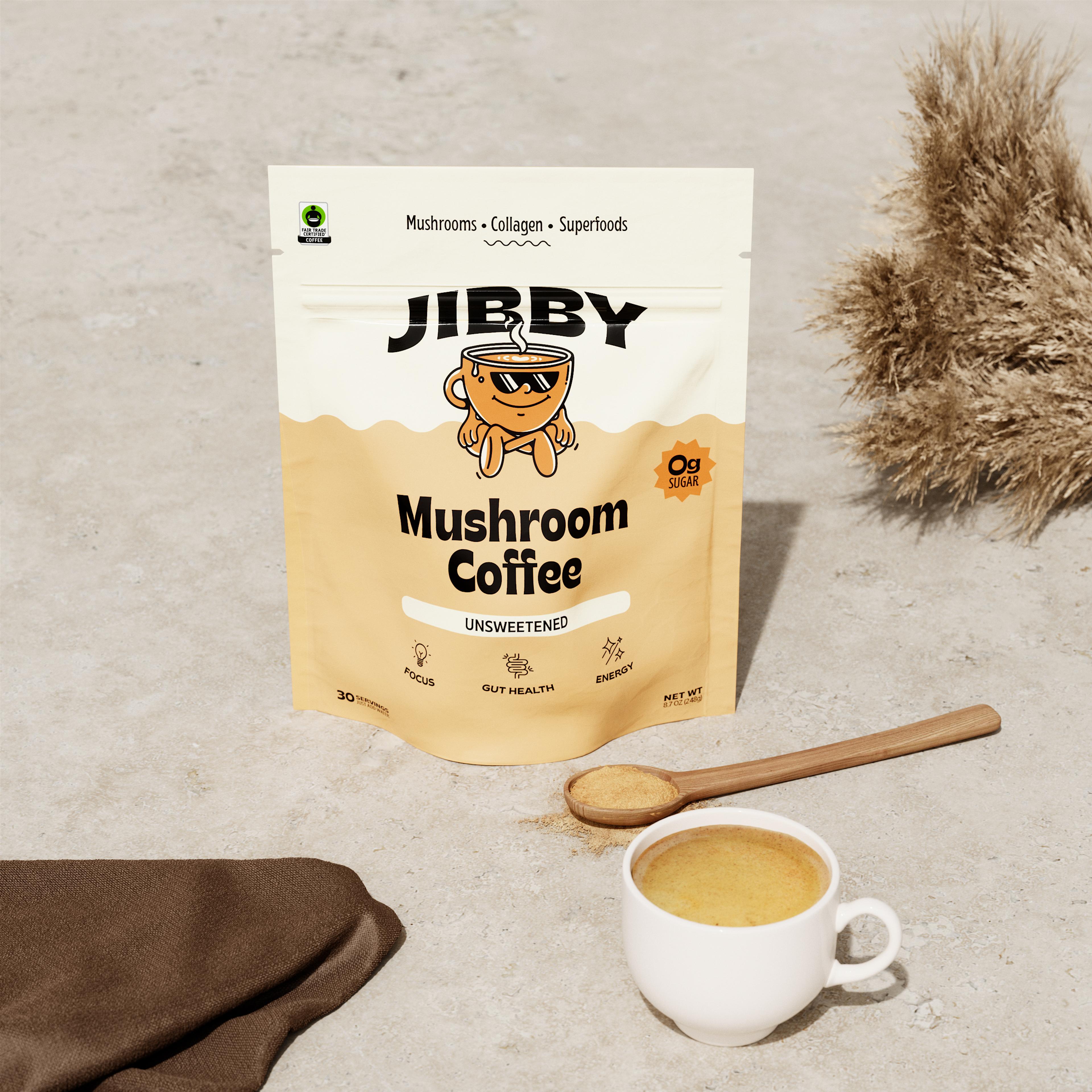 A pouch of Jibby Mushroom Coffee sits on a textured surface with a wooden spoon, a cup of brewed coffee, and dried plants in the background.