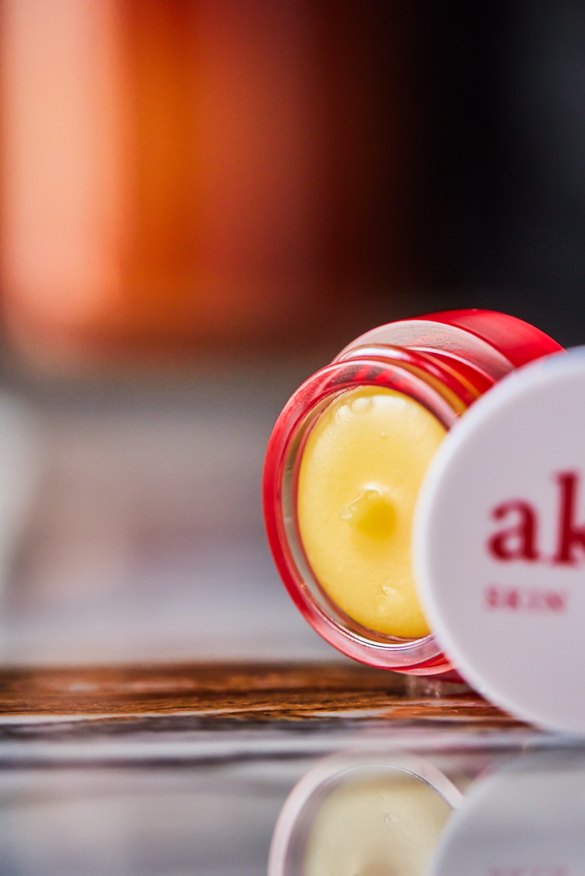 An open Akar skincare balm jar with a red rim, revealing a smooth yellow balm inside.