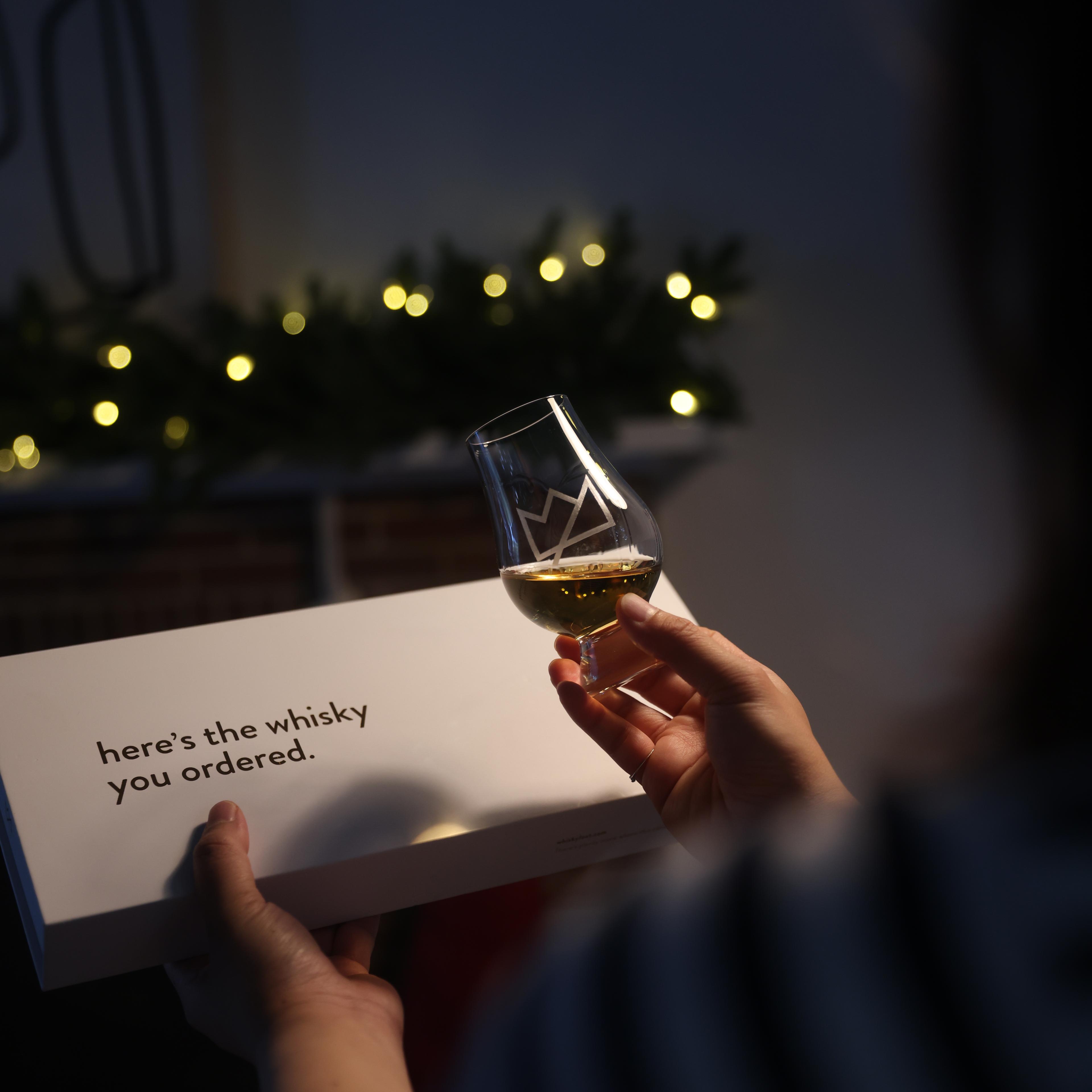 A person holds a whisky tasting glass with golden liquid and a white box labeled "here’s the whisky you ordered," with festive lights glowing in the background.